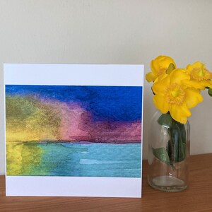May include: A watercolor painting of a sunset over a body of water. The sky is a mix of yellow, pink, purple, and blue. The water is a light blue and green. The painting is on a white card.