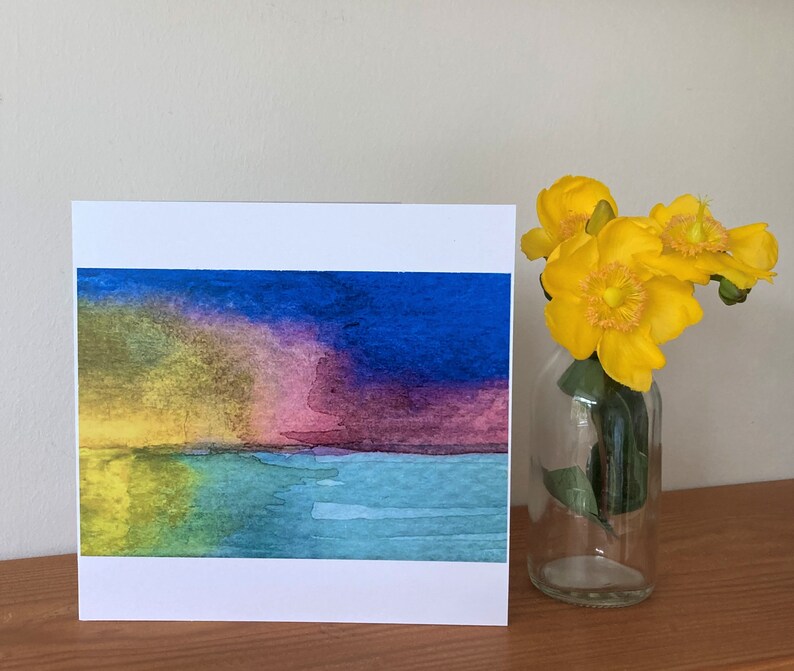 May include: A watercolor painting of a sunset over a body of water. The sky is a mix of yellow, pink, purple, and blue. The water is a light blue and green. The painting is on a white card.