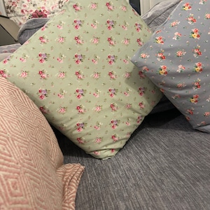 Puede incluir: Tres almohadas decorativas con estampados florales. Las almohadas están sobre un sofá gris. Una almohada es verde con pequeñas flores rosas y blancas, otra es gris con pequeñas flores rosas y amarillas, y la última es rosa con un patrón geométrico.