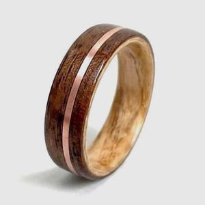 Könnte beinhalten: Ein Ring mit einer dunkelbraunen Holzaußenseite mit sichtbarer Maserung, einer dünnen metallischen kupferfarbenen Einlage, die horizontal um die Mitte verläuft, und einer helleren, glatten Holzauskleidung in Naturtönen.