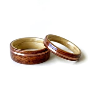 May include: Two rings crafted from dark brown wood with a smooth finish, each featuring a thin, polished copper inlay band running horizontally around the center. The interior of both rings is lined with a lighter, pale-toned wood grain finish.