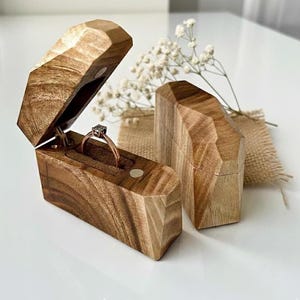 Puede incluir: Un joyero de madera con diseño geométrico, abierto para mostrar un anillo con diamante. La caja es de madera marrón claro y tiene cierre magnético. Hay otra caja cerrada al lado.