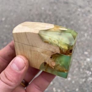 May include: A small wooden box with a hinged lid. The box has a light-coloured wooden top and sides, with a geometric design. The lower section is a translucent green resin with brown inclusions. The box is held in a hand.