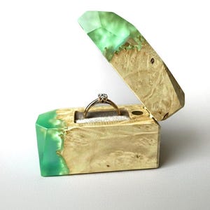 Può includere: Un portaanelli rettangolare realizzato in legno chiaro e resina verde menta traslucida. Il coperchio incernierato è aperto e rivela un interno in velluto grigio con un sottile anello d'oro dotato di una piccola gemma trasparente.