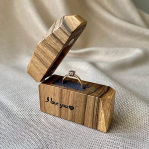 May include: A rectangular wooden ring box with a hinged lid, featuring a natural wood grain pattern and the engraved text "I love you" with a small heart symbol. Inside, a thin gold ring with a small stone sits in a dark blue velvet slot.