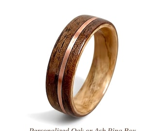 Puede incluir: Un anillo con un exterior de madera marrón oscuro con un patrón de grano visible, una fina incrustación de cobre metálico que recorre horizontalmente el centro y un forro interior de madera lisa de color claro. El texto dice Personalized Oak or Ash Ring Box.