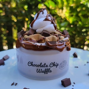 以下が含まれることがあります： チョコレートチップワッフルがホイップクリームとチョコレートソースでトッピングされた白い容器。容器には「チョコレートチップワッフル」の文字が印刷されています。