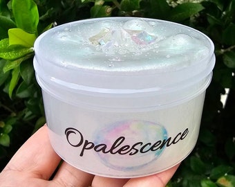 May include: A clear plastic container filled with a white slime with iridescent glitter and a large iridescent crystal. The container has a black label with the word "Opalescence" written in a cursive font.