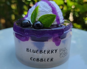 May include: A miniature blueberry cobbler dessert with a white frosting topping and purple blueberry sauce. The dessert is in a clear plastic container with the words "Blueberry Cobbler" printed on the side.
