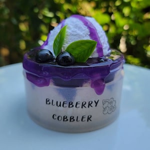 May include: A miniature blueberry cobbler dessert with a white frosting topping and purple blueberry sauce. The dessert is in a clear plastic container with the words "Blueberry Cobbler" printed on the side.
