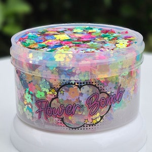 May include: A clear plastic container filled with colourful iridescent flower-shaped glitter. The container has a white lid and a label that reads "Flower Bomb".
