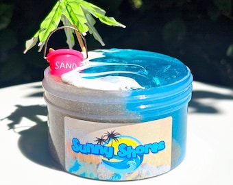 May include: A clear plastic jar filled with blue and gray slime. The slime is topped with a white wave and a pink bucket with the word "SAND" on it. A green palm tree umbrella sits on top of the bucket. The jar has a label that says "Sunny Shores".