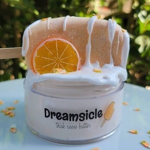 May include: A white jar of orange-flavored "Dreamsicle" thick snow butter with a white and orange popsicle on top. The jar is decorated with a white and orange popsicle graphic and the text "Dreamsicle thick snow butter".