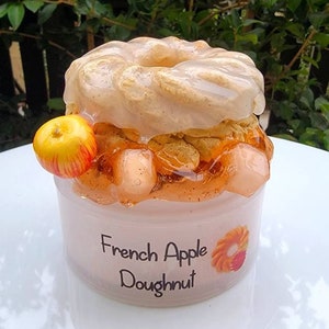 May include: A clear glass jar filled with a layered dessert. The bottom layer is a light brown color, the middle layer is a light orange color, and the top layer is a white donut with a glaze. The jar is labeled "French Apple Doughnut".