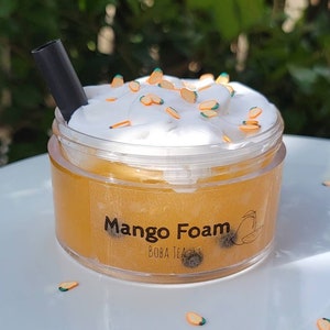K&ouml;nnte beinhalten: Ein durchsichtiger Plastikbecher gef&uuml;llt mit einer gelben Fl&uuml;ssigkeit, bedeckt mit wei&szlig;er Schlagsahne und orangefarbenen Streuseln. Der Becher hat ein schwarzes Etikett mit der Aufschrift "Mango Foam Boba Tea".