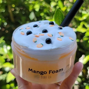 K&ouml;nnte beinhalten: Ein durchsichtiger Plastikbecher gef&uuml;llt mit einer gelben Fl&uuml;ssigkeit, bedeckt mit wei&szlig;er Schlagsahne und schwarzen Boba-Perlen. Der Becher tr&auml;gt ein Etikett mit der Aufschrift "Mango Foam Boba Tea".