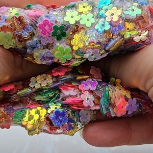 May include: A clear slime with iridescent flower-shaped sequins in various colours, including pink, blue, green, yellow, and orange. The slime is being held in two hands.