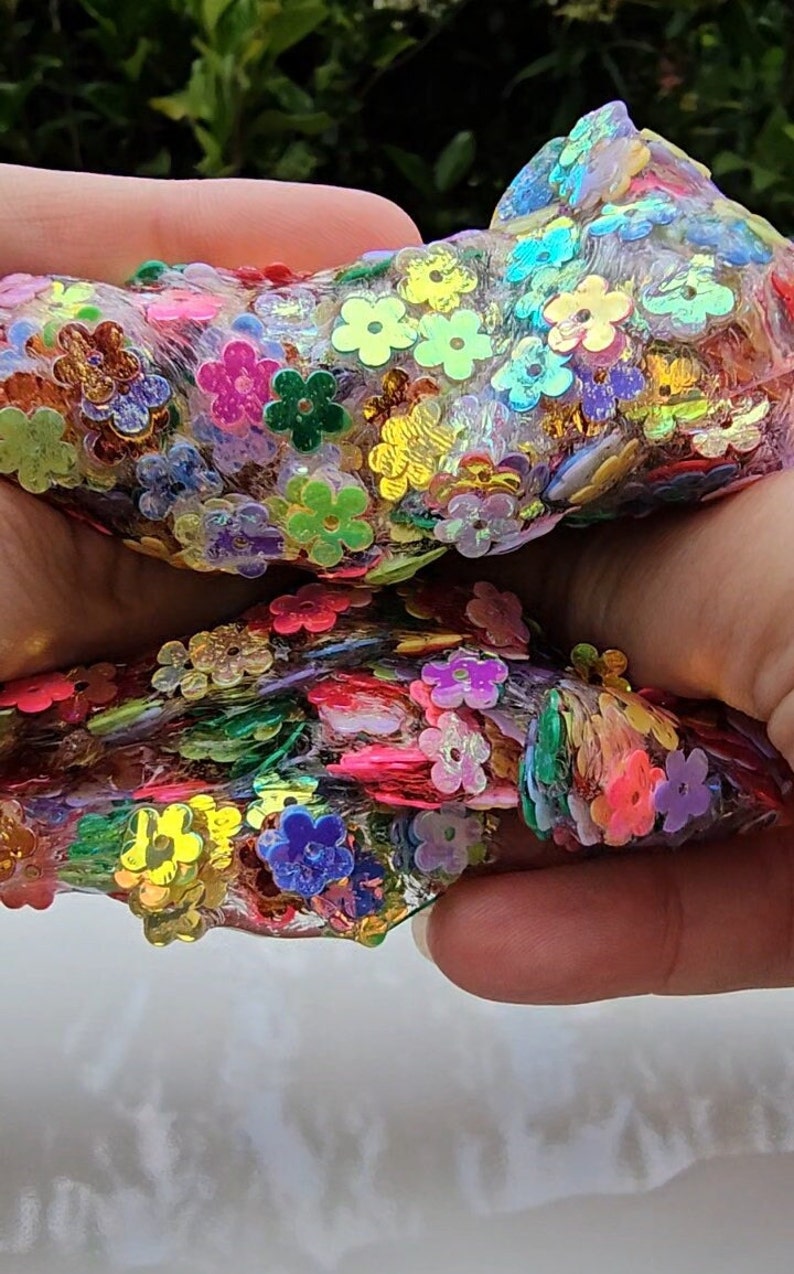 May include: A clear slime with iridescent flower-shaped sequins in various colours, including pink, blue, green, yellow, and orange. The slime is being held in two hands.