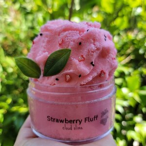 Peut inclure: Slime rose en nuage dans un pot transparent avec une &eacute;tiquette noire indiquant "Strawberry Fluff cloud slime". Le slime est surmont&eacute; de paillettes noires et rouges et de feuilles vertes.