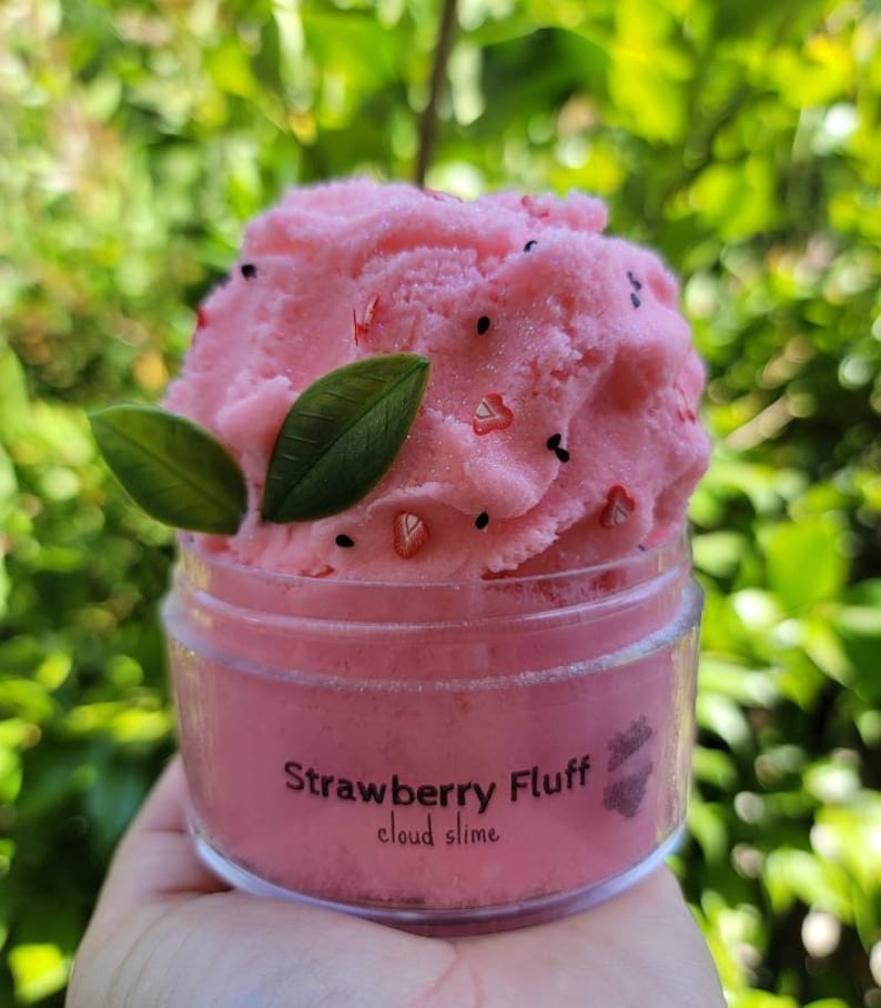 Peut inclure: Slime rose en nuage dans un pot transparent avec une &eacute;tiquette noire indiquant "Strawberry Fluff cloud slime". Le slime est surmont&eacute; de paillettes noires et rouges et de feuilles vertes.