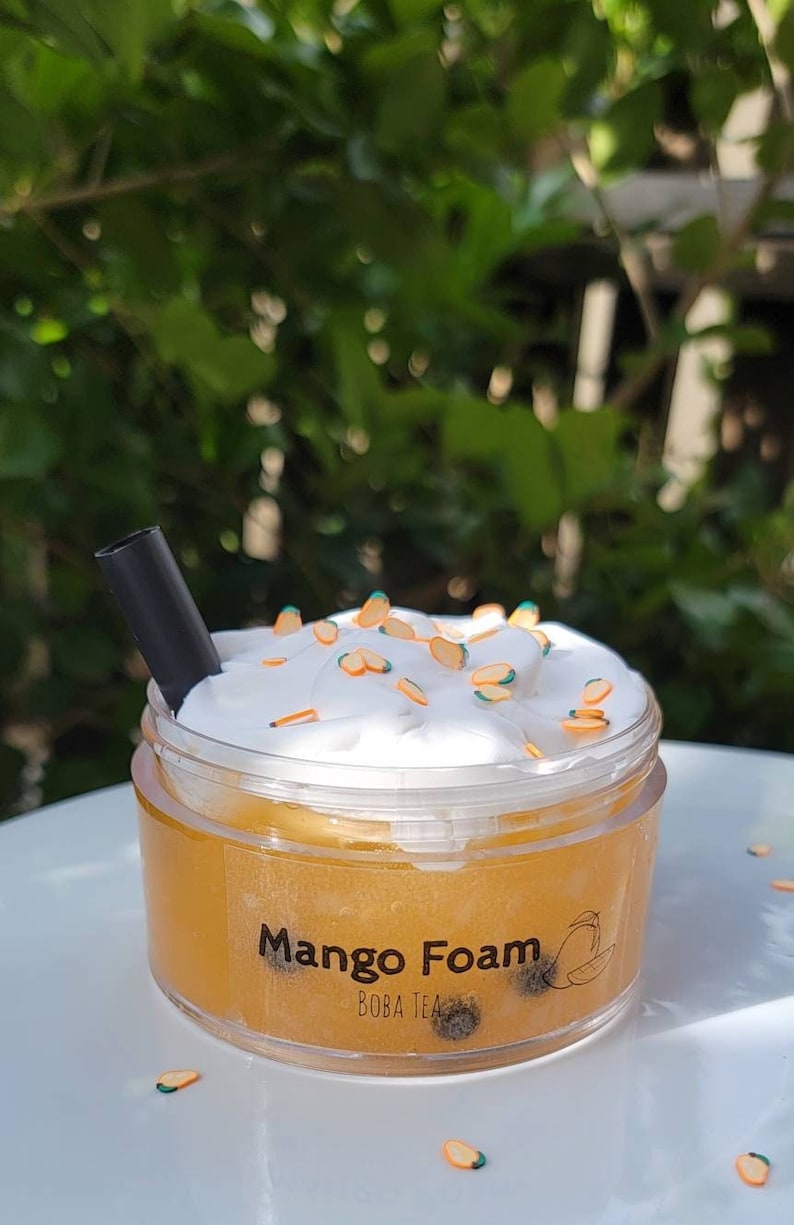 K&ouml;nnte beinhalten: Ein durchsichtiger Plastikbecher gef&uuml;llt mit einer gelben Fl&uuml;ssigkeit, bedeckt mit wei&szlig;er Schlagsahne und orangefarbenen Streuseln. Der Becher hat ein schwarzes Etikett mit der Aufschrift "Mango Foam Boba Tea".