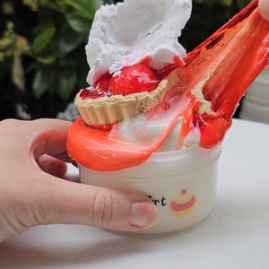 May include: A red and white mini pie with whipped cream topping is being poured into a white container with a red slime. The container has a label that says "Tart" and a small cupcake graphic.