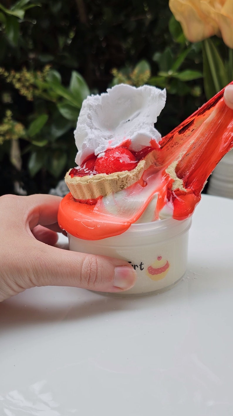 May include: A red and white mini pie with whipped cream topping is being poured into a white container with a red slime. The container has a label that says "Tart" and a small cupcake graphic.