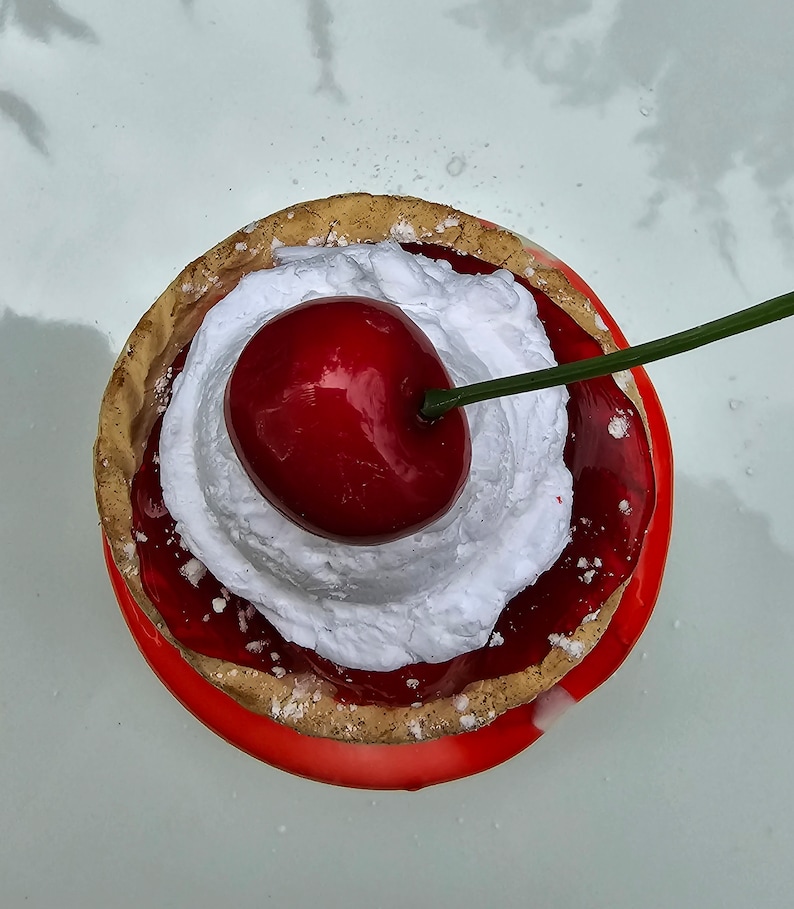 May include: A red cherry with a green stem sits on top of a white whipped cream topping on a cherry pie. The pie is in a red and brown pastry crust.