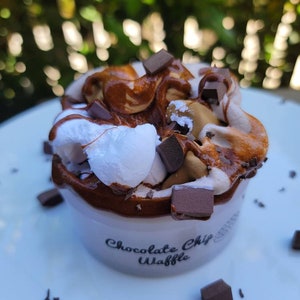 以下が含まれることがあります： 白いセラミックカップにチョコレートチップワッフル生地が満たされ、ホイップクリームとチョコレートチップがトッピングされています。カップは白いプレートの上にあり、周りにチョコレートチップが散らばっています。