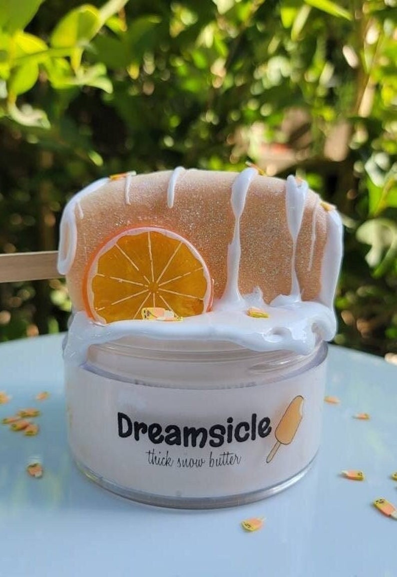 May include: A white jar of orange-flavored "Dreamsicle" thick snow butter with a white and orange popsicle on top. The jar is decorated with a white and orange popsicle graphic and the text "Dreamsicle thick snow butter".