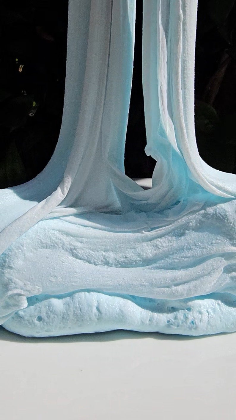 May include: A close-up of light blue slime, stretched and pulled into a long, thin shape. The slime is soft and gooey, with a slightly textured surface.