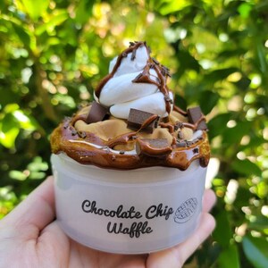 以下が含まれることがあります： チョコレートチップワッフルのスライムが入った白い容器。スライムはホイップクリームとチョコレートソースでトッピングされています。容器には「チョコレートチップワッフル」という文字が印刷されています。