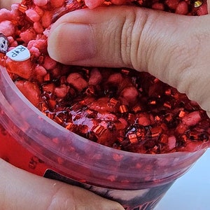 May include: A red slime with red glitter in a clear plastic container. The slime is being squeezed by a hand. There are small red and black stickers on a white surface.