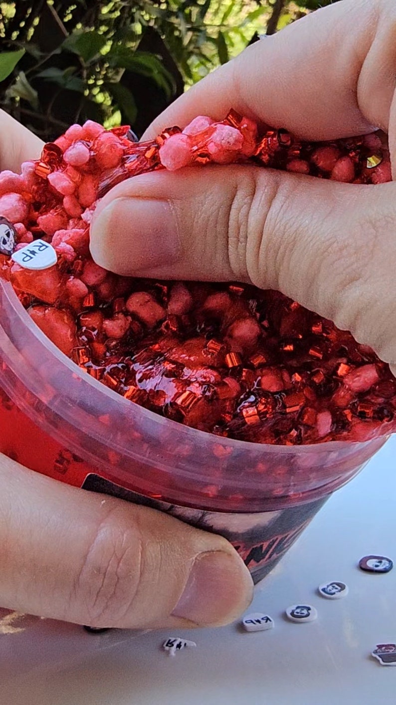 May include: A red slime with red glitter in a clear plastic container. The slime is being squeezed by a hand. There are small red and black stickers on a white surface.