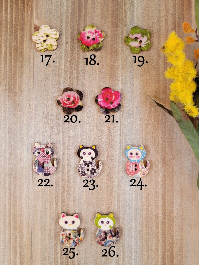 Op de afbeelding: Assortiment van 26 houten knopen met verschillende bloemen- en dierenmotieven. De knopen zijn in tinten roze, groen, blauw, wit en zwart. De knopen zijn genummerd van 17 tot en met 26.