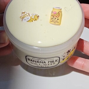 May include: A clear plastic container filled with a light yellow slime. The container has a label that reads "Crafty Farms Banana Milk Grade A | Vit A & D Do Not Eat". There are small decorative charms on top of the slime, including a yellow cow and a yellow can with Japanese text that reads "バナナミルク クリーミー".
