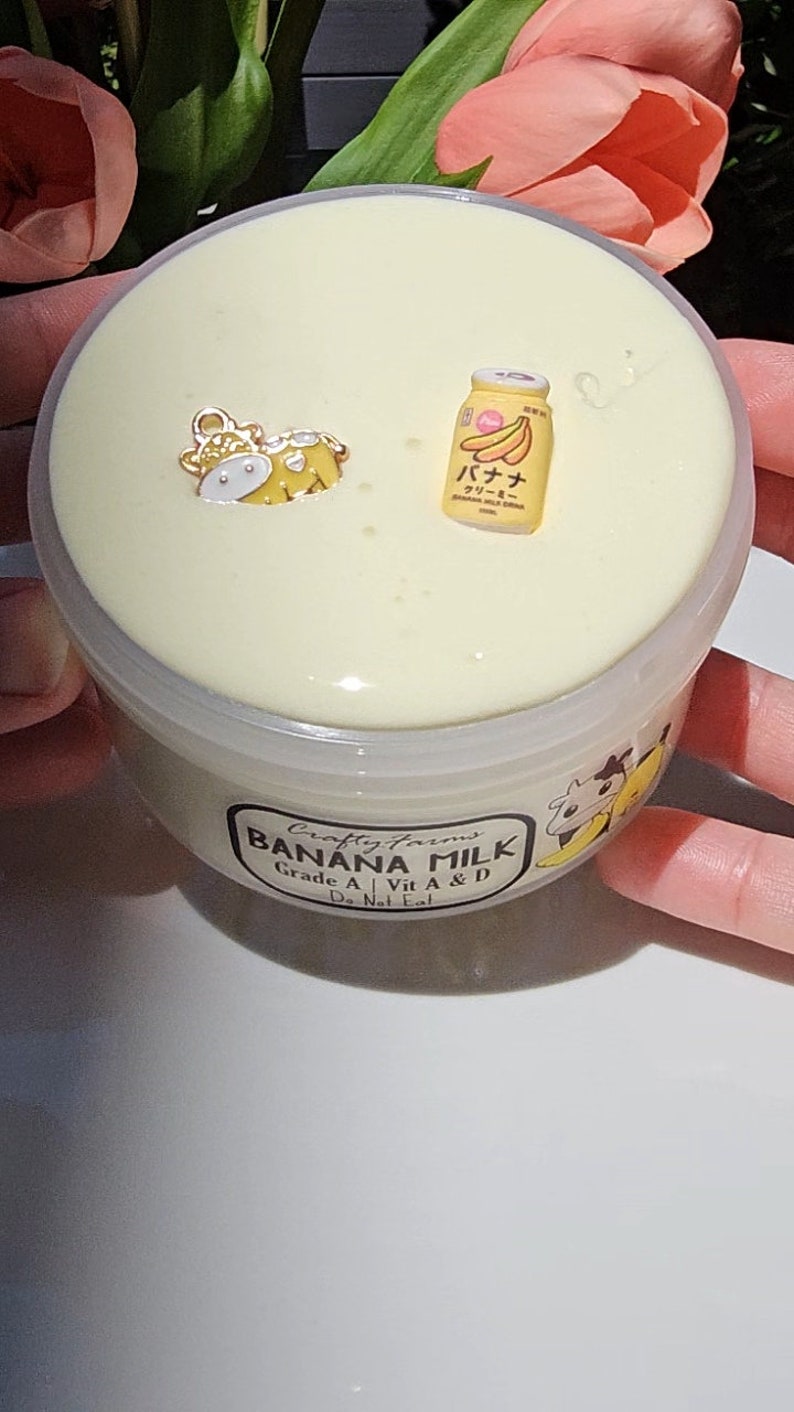 May include: A clear plastic container filled with a light yellow slime. The container has a label that reads "Crafty Farms Banana Milk Grade A | Vit A & D Do Not Eat". There are small decorative charms on top of the slime, including a yellow cow and a yellow can with Japanese text that reads "バナナミルク クリーミー".