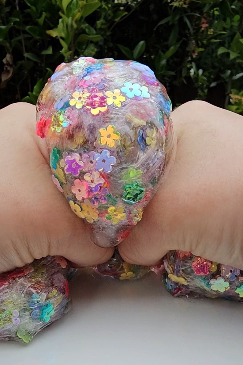 May include: A clear slime with iridescent flower-shaped glitter embedded throughout. The slime is being held in two hands.