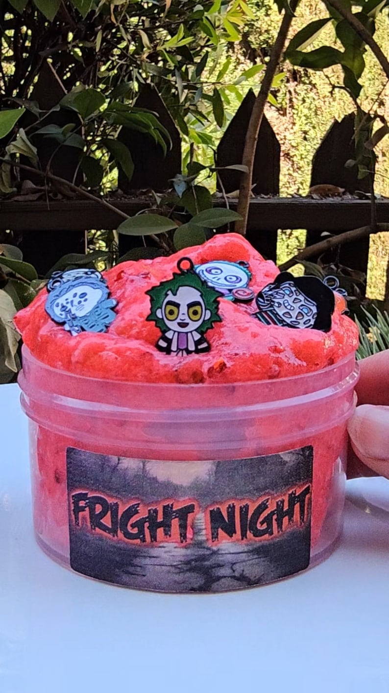 May include: A red slime with a black and white label that says "FRIGHT NIGHT". The slime has several small charms in it, including a cartoon character with green hair and a black and white face.