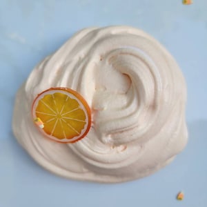 May include: A close-up of a light pink slime with an orange citrus slice on top. The slime is on a white plate.