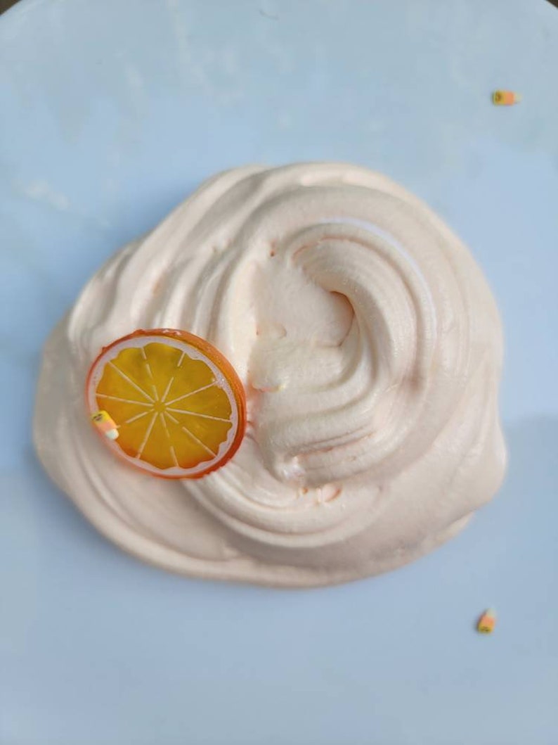 May include: A close-up of a light pink slime with an orange citrus slice on top. The slime is on a white plate.