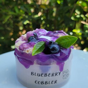 May include: A small clear plastic container with a purple blueberry cobbler inside. The cobbler is topped with blueberries and a few green leaves. The container has the words "Blueberry Cobbler" printed on the side.