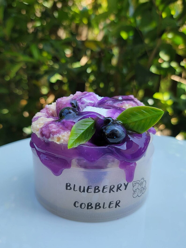 May include: A small clear plastic container with a purple blueberry cobbler inside. The cobbler is topped with blueberries and a few green leaves. The container has the words "Blueberry Cobbler" printed on the side.