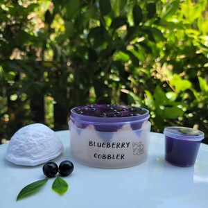 May include: A clear plastic container filled with a purple blueberry cobbler dessert topped with blueberries. The container has a white label that reads "Blueberry Cobbler". There are two small blueberries and a small white ball of dough on a white surface.