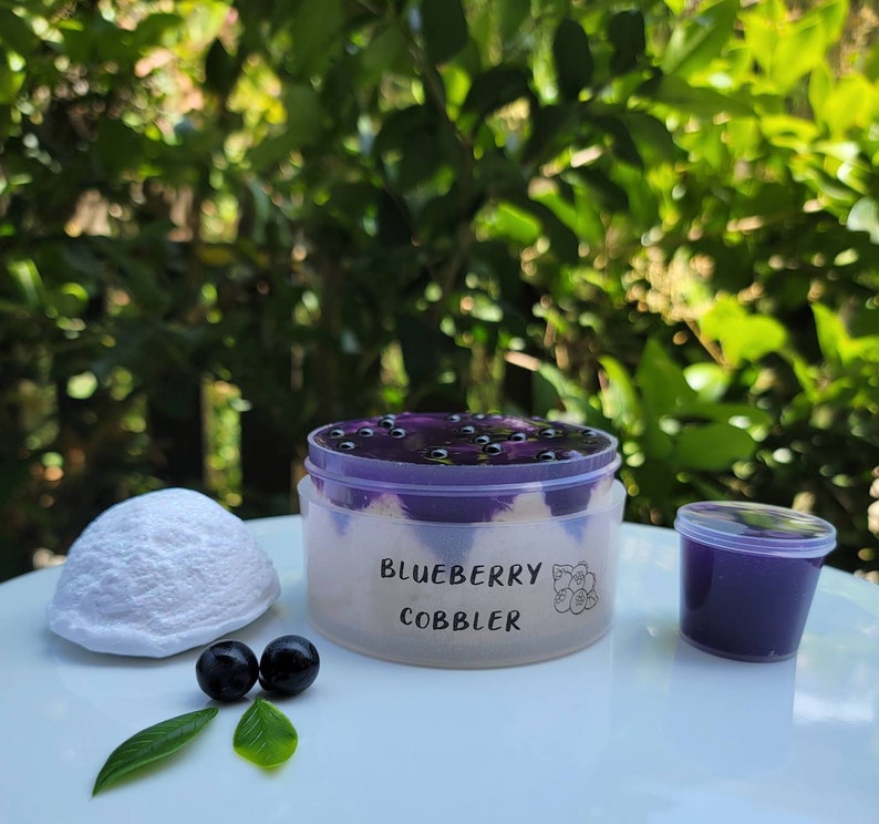 May include: A clear plastic container filled with a purple blueberry cobbler dessert topped with blueberries. The container has a white label that reads "Blueberry Cobbler". There are two small blueberries and a small white ball of dough on a white surface.