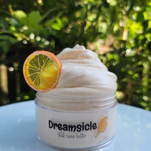 May include: A clear jar of thick snow butter with a white and orange label that says "Dreamsicle thick snow butter". A slice of orange is on top of the jar.