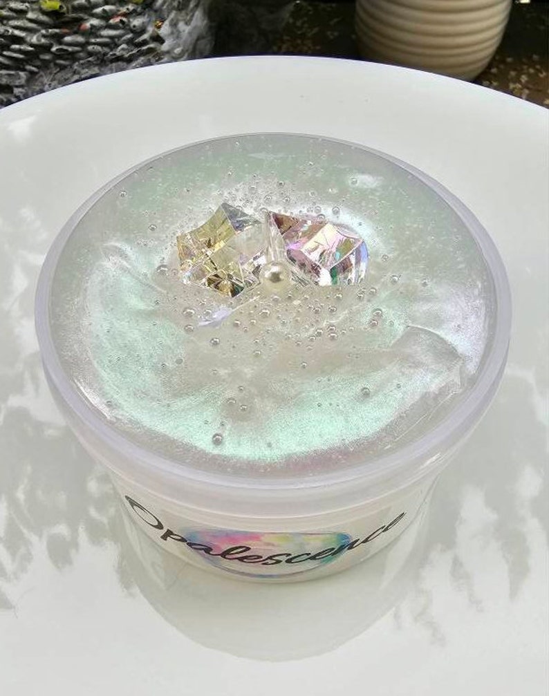 Op de afbeelding: Een doorzichtige container met iriserende slijm met twee iriserende kristallen en een parel erin. De container heeft een wit label met het woord "Opalescence" erop gedrukt.