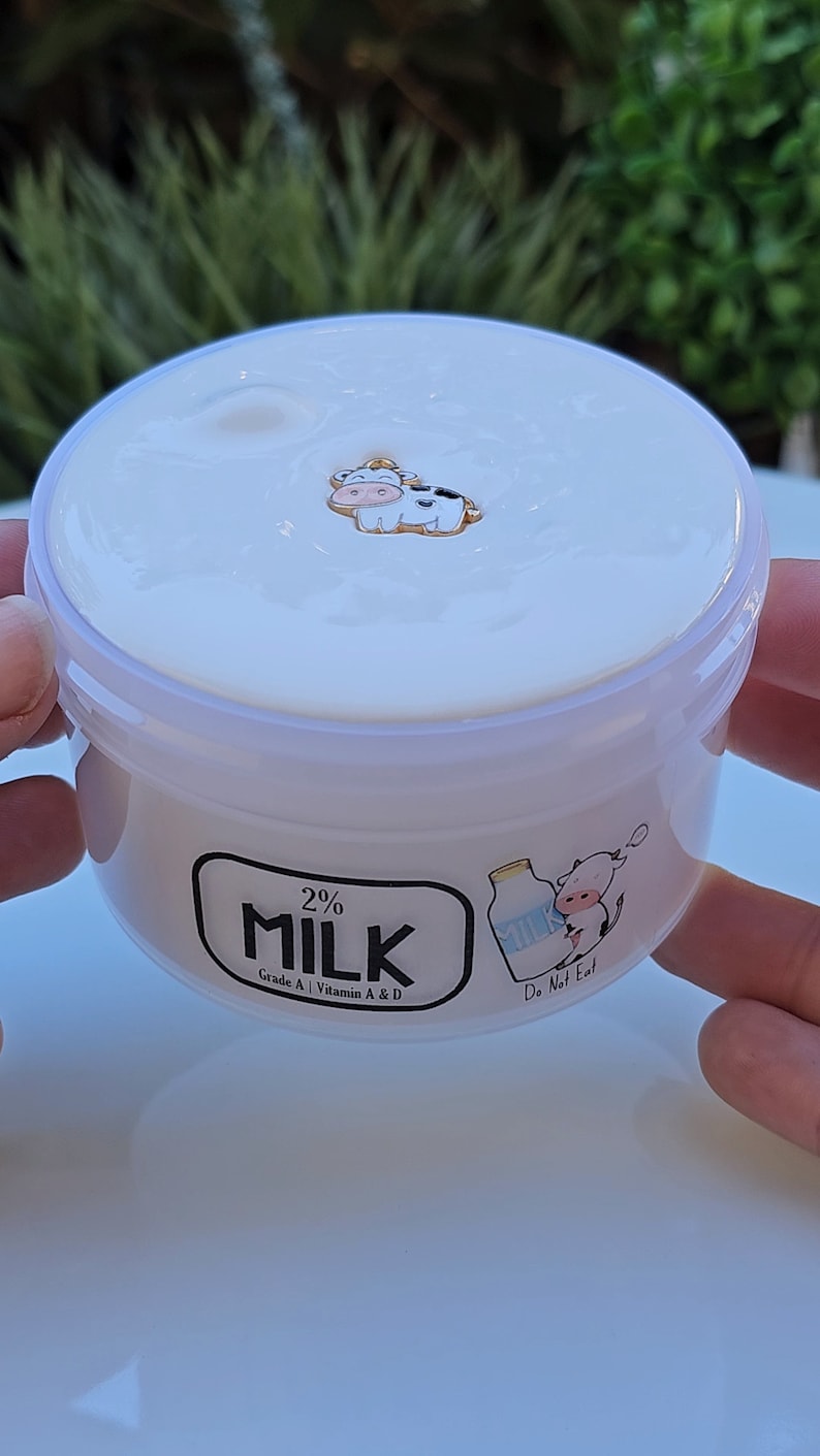 May include: A white plastic container with a white lid filled with a white substance. The container has a black label that says "2% MILK Grade A | Vitamin A & D" and a cartoon cow with a milk carton. The label also says "Do Not Eat".