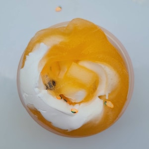 K&ouml;nnte beinhalten: Nahaufnahme eines durchsichtigen Beh&auml;lters, gef&uuml;llt mit einem Wirbel aus gelbem und wei&szlig;em Schleim mit orangefarbenem und gr&uuml;nem Glitzer.