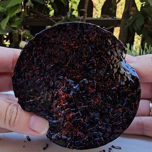 K&ouml;nnte beinhalten: Ein runder Untersetzer aus schwarzem und rotem Glitzerharz. Der Untersetzer ist mit kleinen schwarzen und roten Glitzerst&uuml;cken bedeckt.